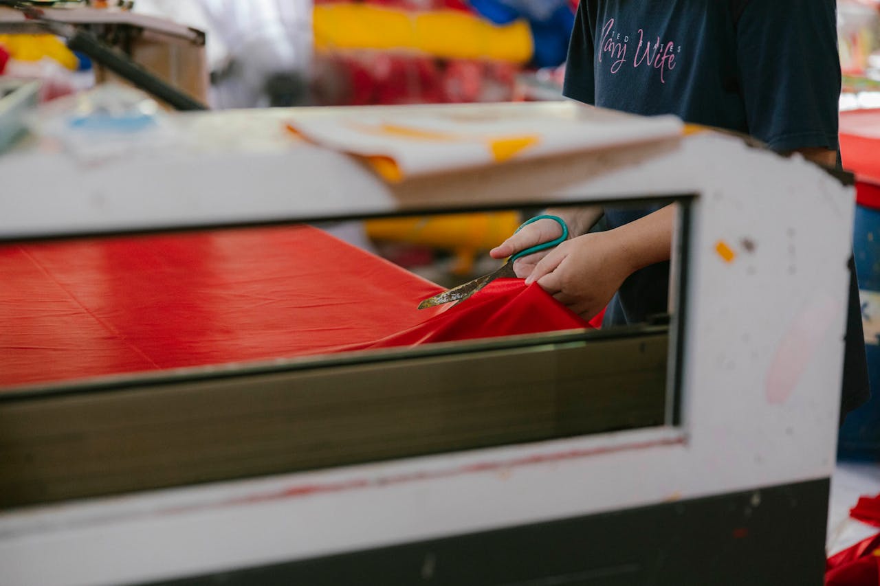 A factory worker precisely cuts red fabric with scissors, showcasing textile production.